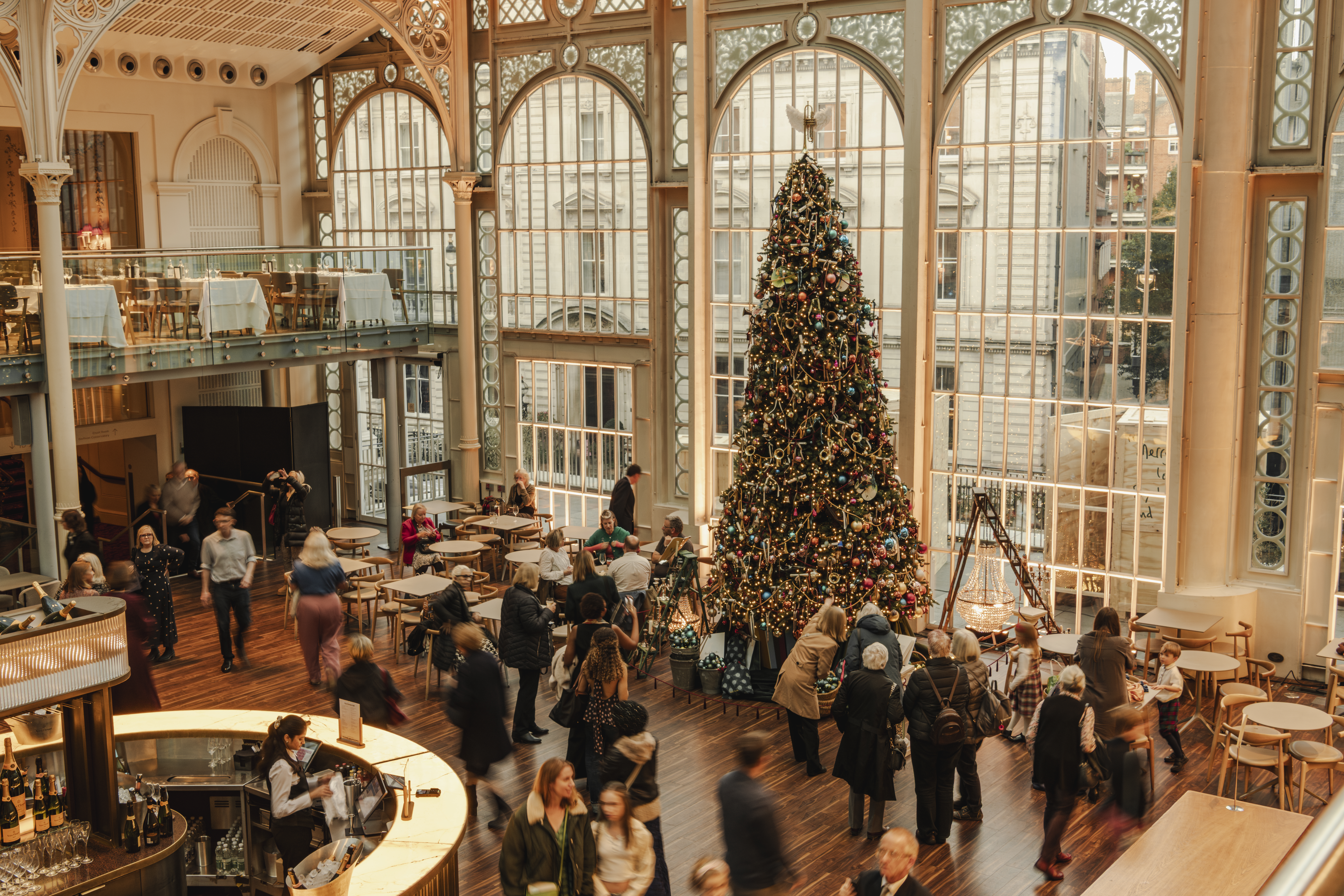 Spacious hall with tall arched windows, filled with people socialising. A large, decorated Christmas tree stands prominently, creating a festive atmosphere. (1 of 3)