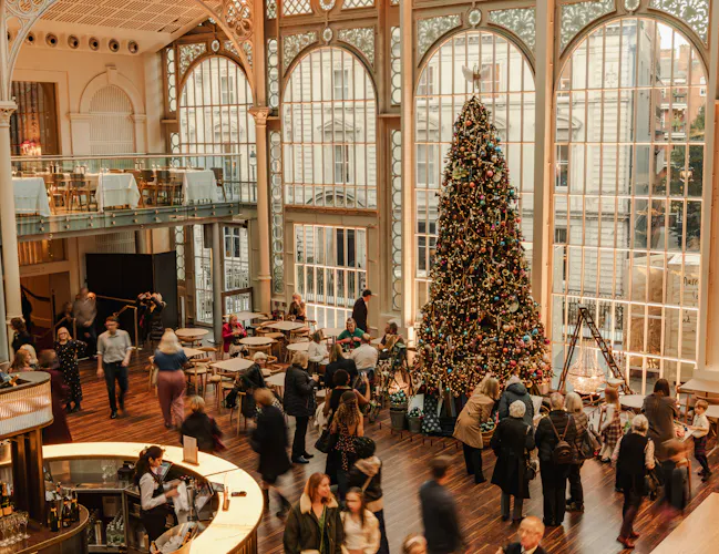 Spacious hall with tall arched windows, filled with people socialising. A large, decorated Christmas tree stands prominently, creating a festive atmosphere. (1 of 3)