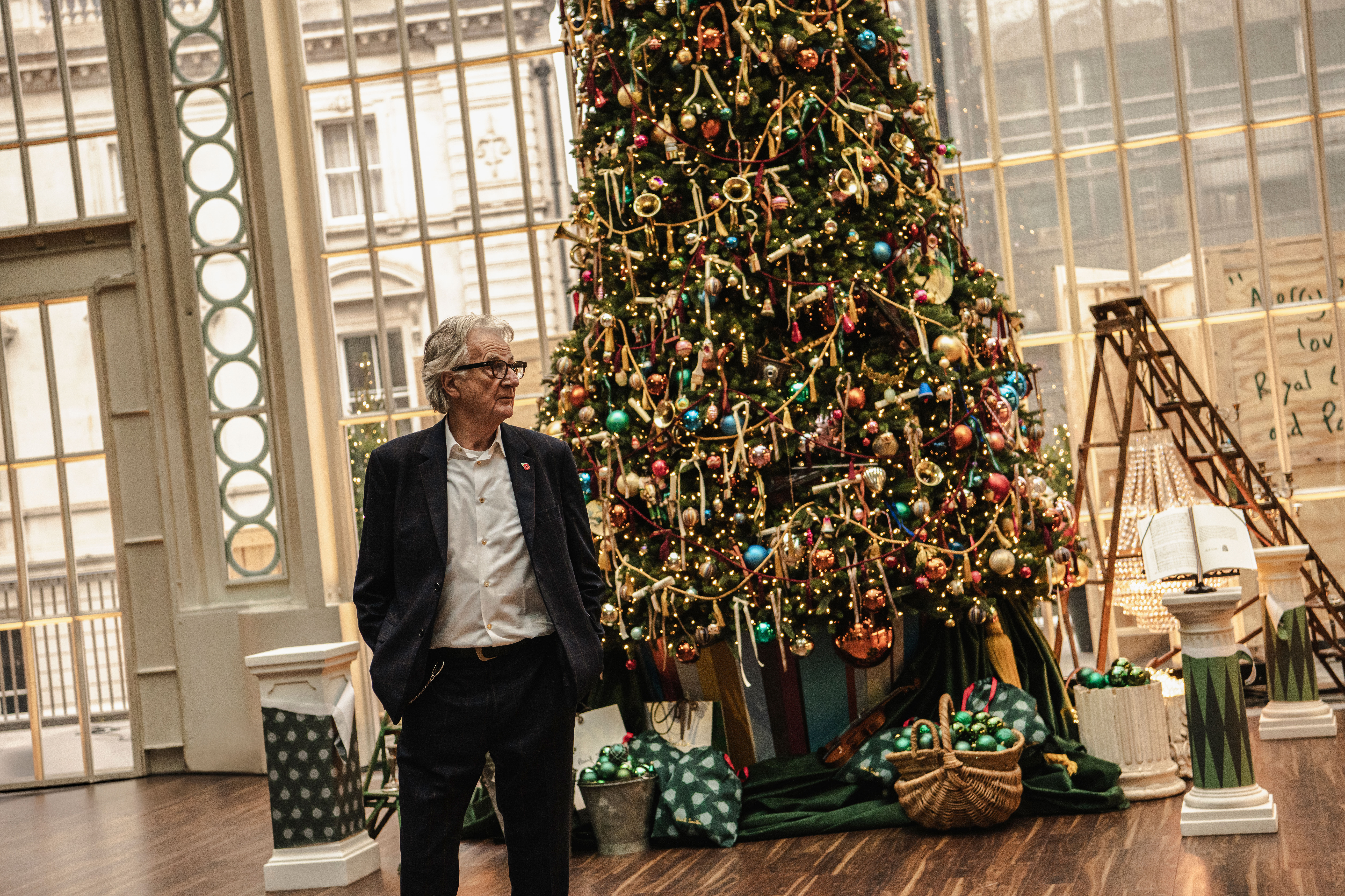 A man in a navy suit stands beside a large, elaborately decorated Christmas tree indoors. 