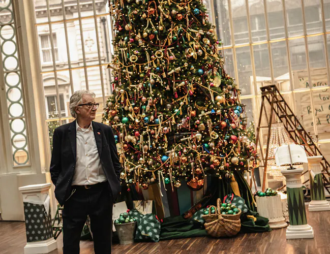 A man in a navy suit stands beside a large, elaborately decorated Christmas tree indoors. (2 of 3)