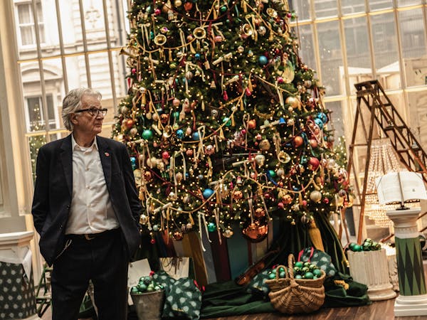 A man in a navy suit stands beside a large, elaborately decorated Christmas tree indoors.