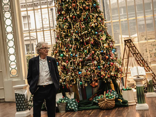A man in a navy suit stands beside a large, elaborately decorated Christmas tree indoors.