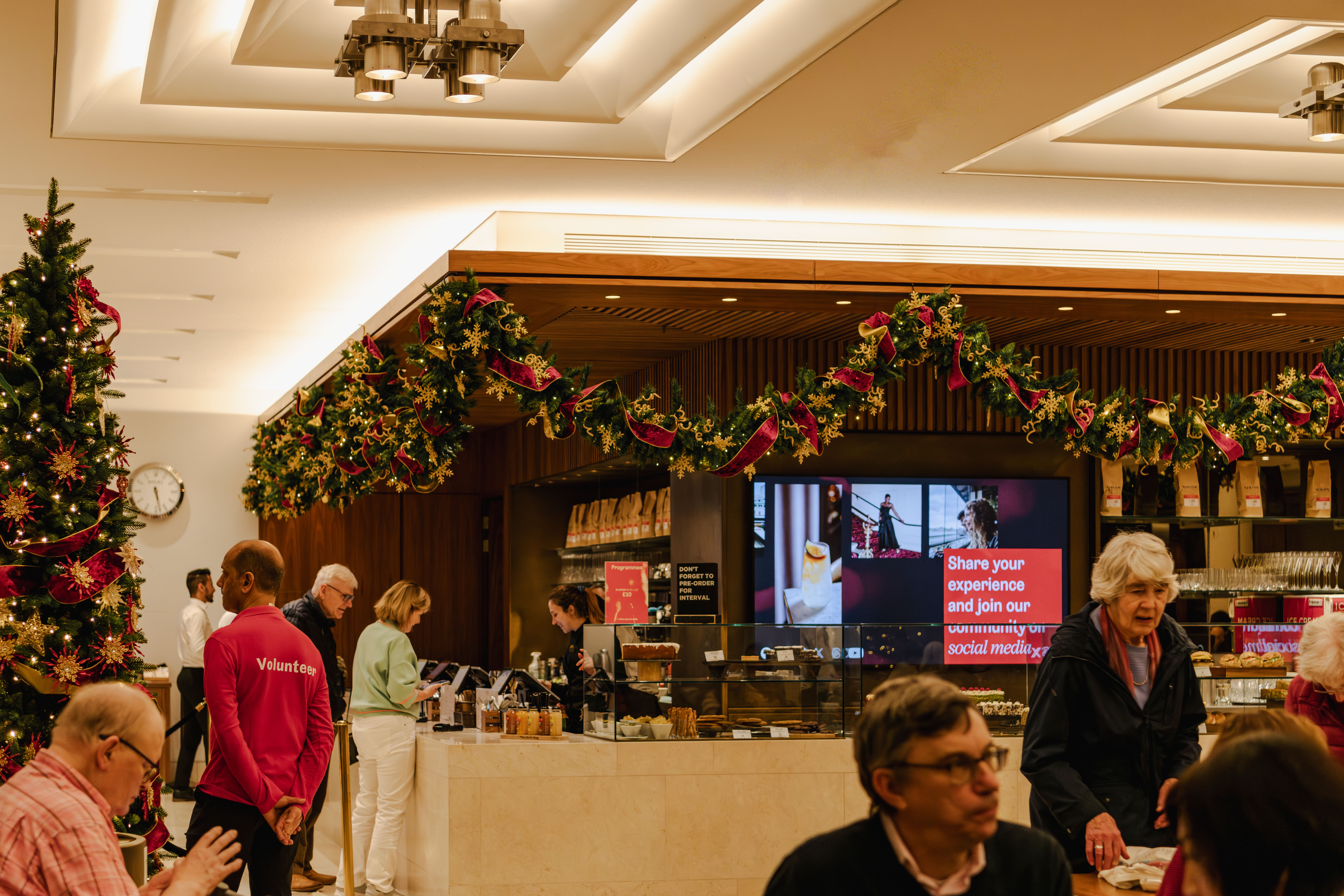 An indoor café decorated with Christmas garlands and a tree. People order at the counter, looking at cakes in glass cases. 