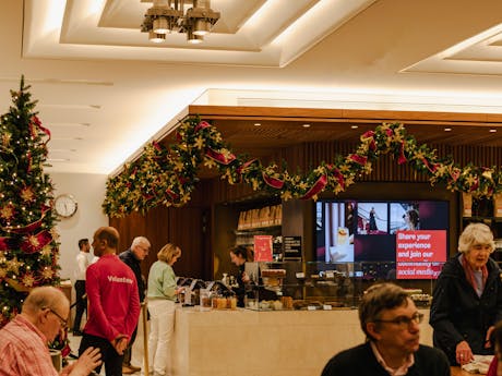 An indoor café decorated with Christmas garlands and a tree. People order at the counter, looking at cakes in glass cases.
