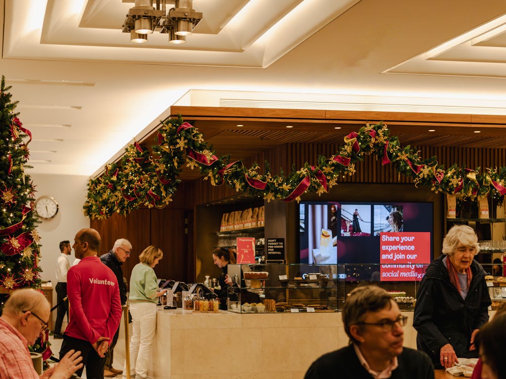 An indoor café decorated with Christmas garlands and a tree. People order at the counter, looking at cakes in glass cases.