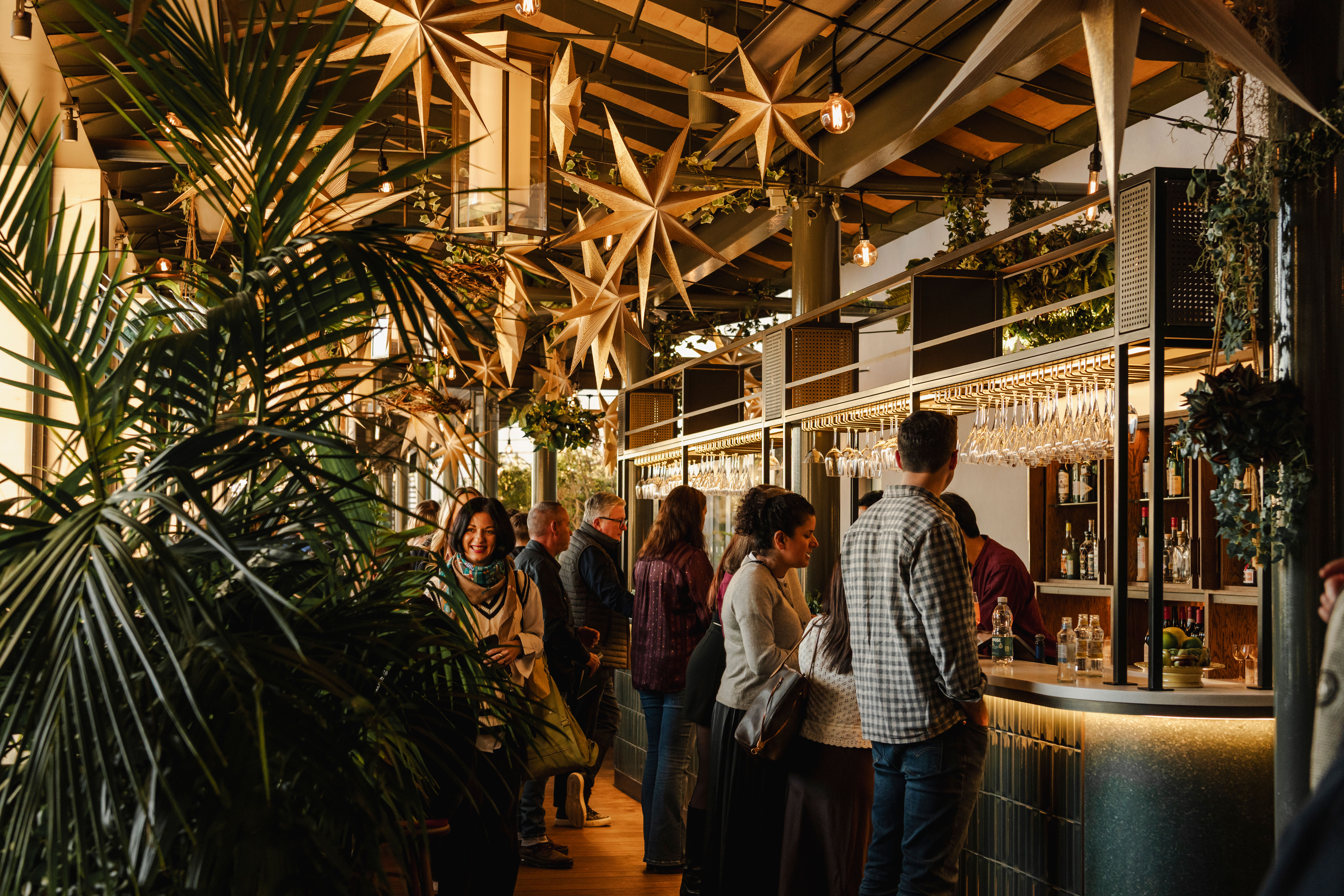A lively bar scene with people socializing. Golden star decorations hang from the ceiling, and lush greenery surrounds the area.