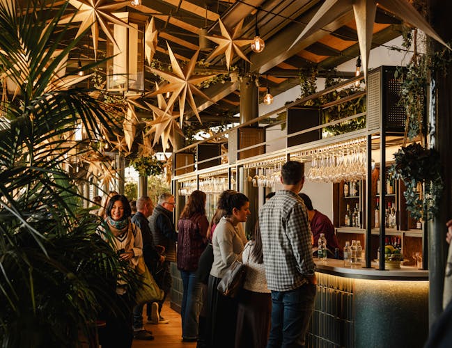A lively bar scene with people socializing. Golden star decorations hang from the ceiling, and lush greenery surrounds the area.