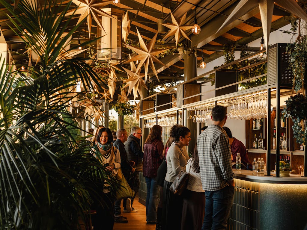 A lively bar scene with people socializing. Golden star decorations hang from the ceiling, and lush greenery surrounds the area.