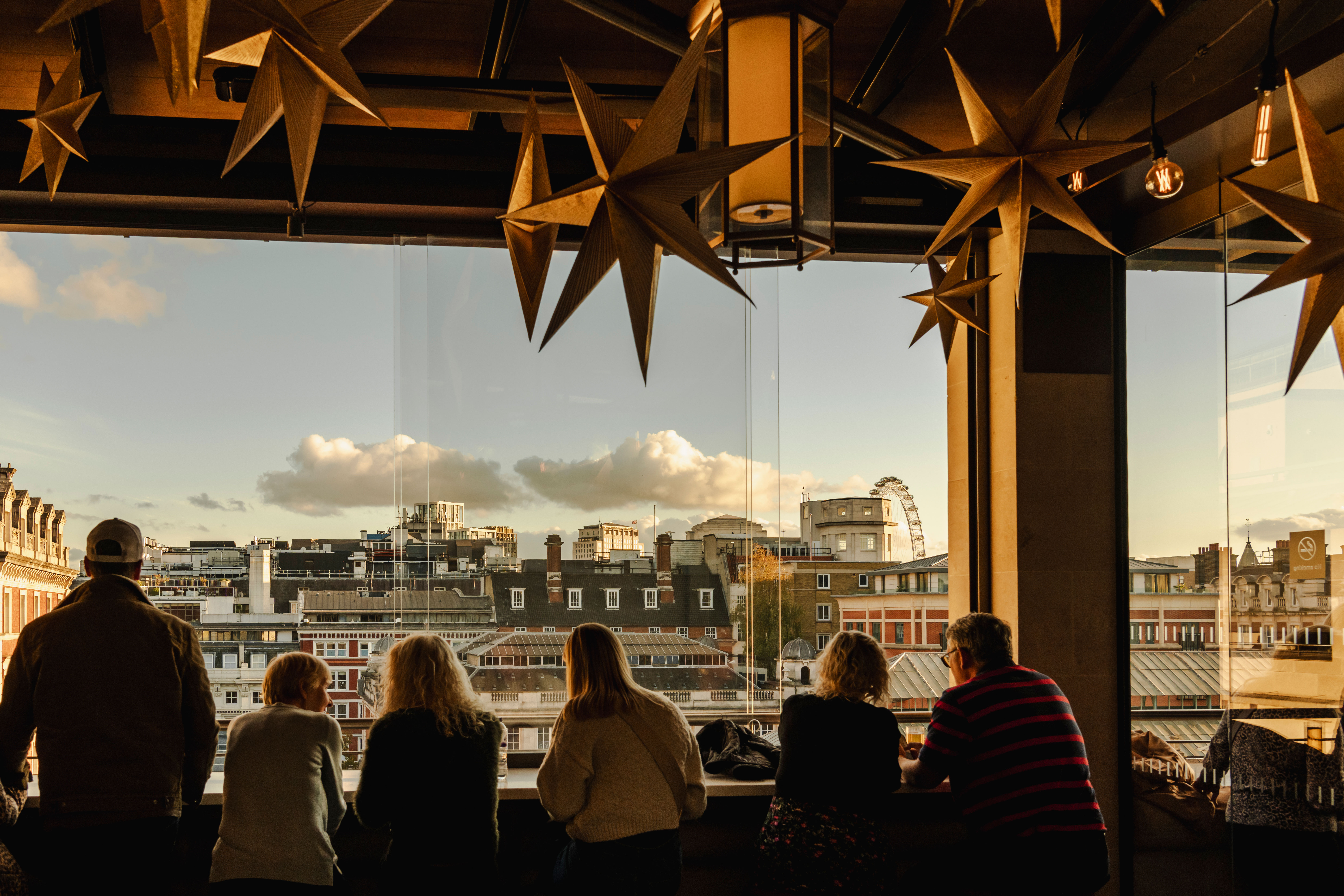 Silhouetted group of people gaze out from a bar, decorated with hanging star ornaments, overlooking the London skyline under a cloudy blue sky.
