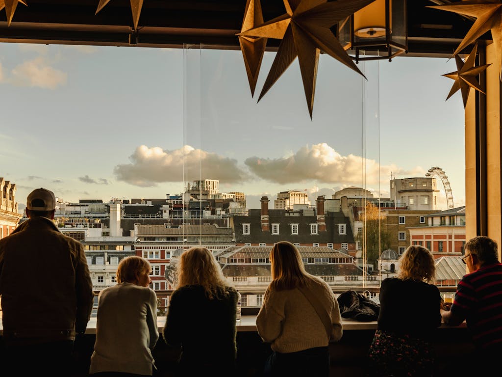 Silhouetted group of people gaze out from a bar, decorated with hanging star ornaments, overlooking the London skyline under a cloudy blue sky.