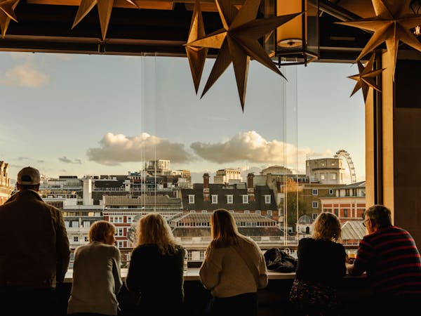 Silhouetted group of people gaze out from a bar, decorated with hanging star ornaments, overlooking the London skyline under a cloudy blue sky.