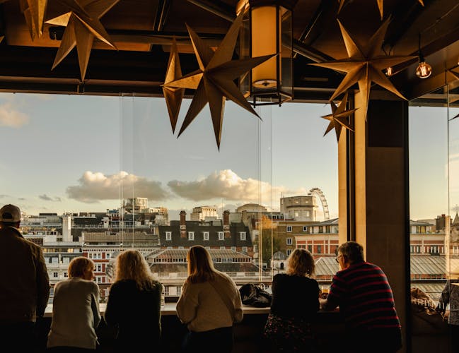 Silhouetted group of people gaze out from a bar, decorated with hanging star ornaments, overlooking the London skyline under a cloudy blue sky.