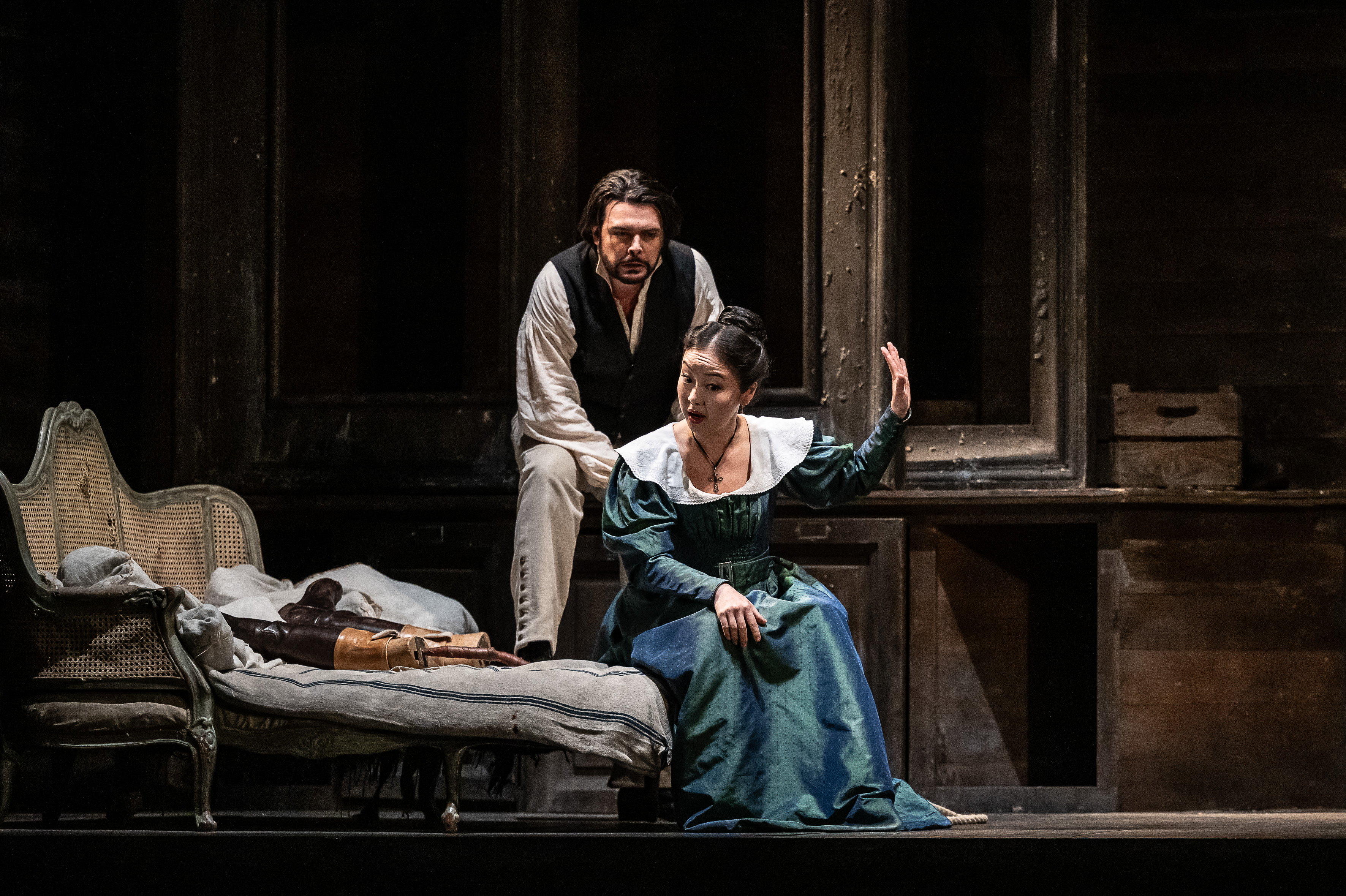 A woman in a blue dress holds up her hand to silence a man in a white shirt and dark waistcoat who is trying to talk to her. She is sat on a chair. 
