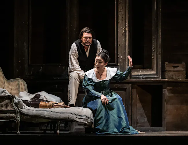 A woman in a blue dress holds up her hand to silence a man in a white shirt and dark waistcoat who is trying to talk to her. She is sat on a chair.