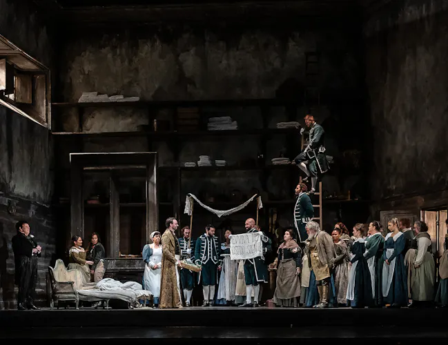 A group of servants are in a dark room being decorated for a wedding. One of them holds a sign