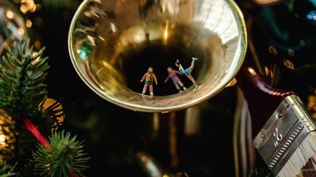 Tiny figures dancing inside a golden trumpet bell, hung on a festive Christmas tree with blurred green needles and colourful decorations.