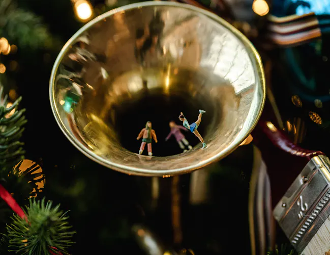 Tiny figures dancing inside a golden trumpet bell, hung on a festive Christmas tree with blurred green needles and colourful decorations. (3 of 3)