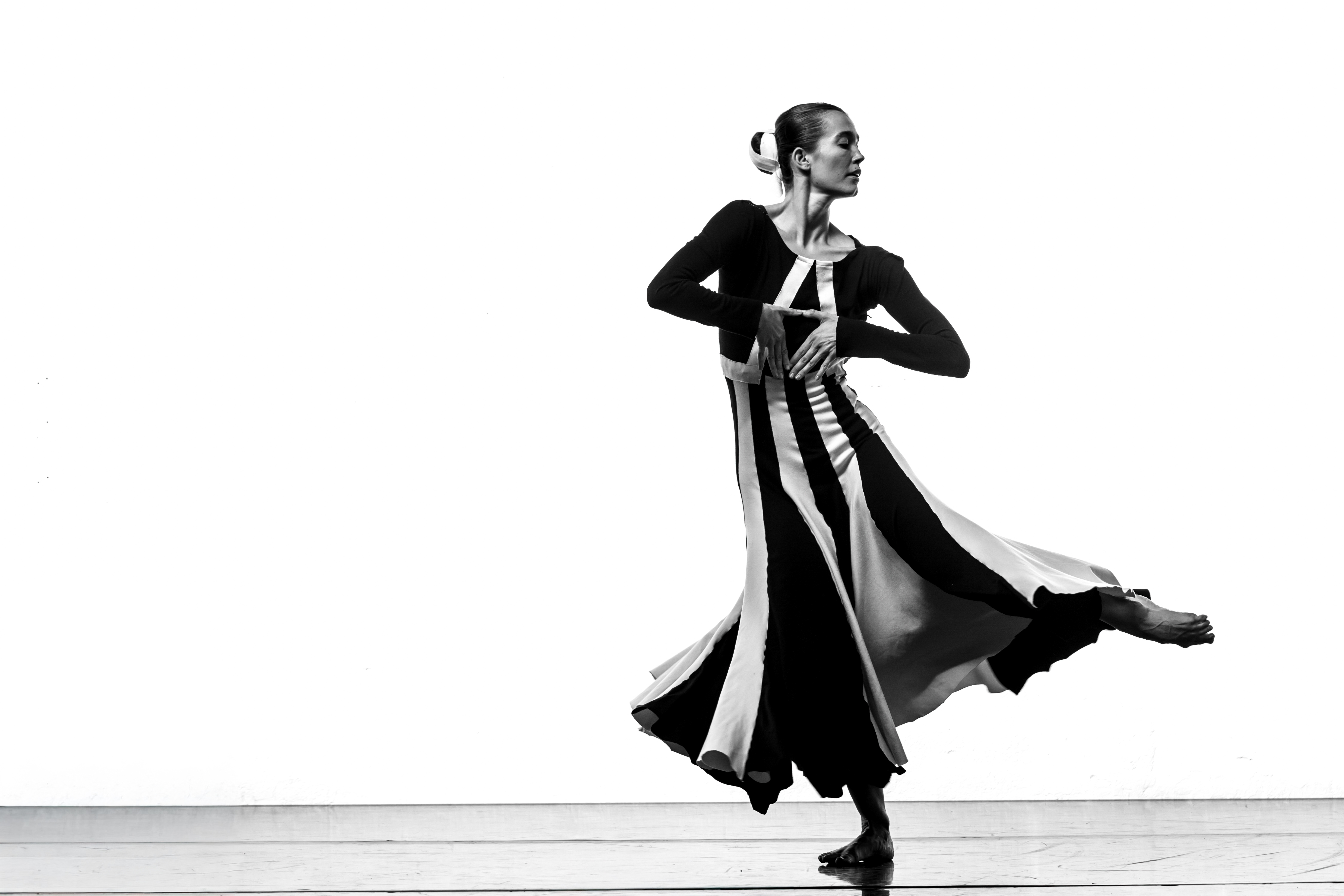 A dancer from Yorke Dance Company in a flowing, striped dress performs a graceful pose on stage, captured in black and white. She stands on one foot with the other leg extended to the side, arms curved in front of her body, and her hair pulled back in a bun.