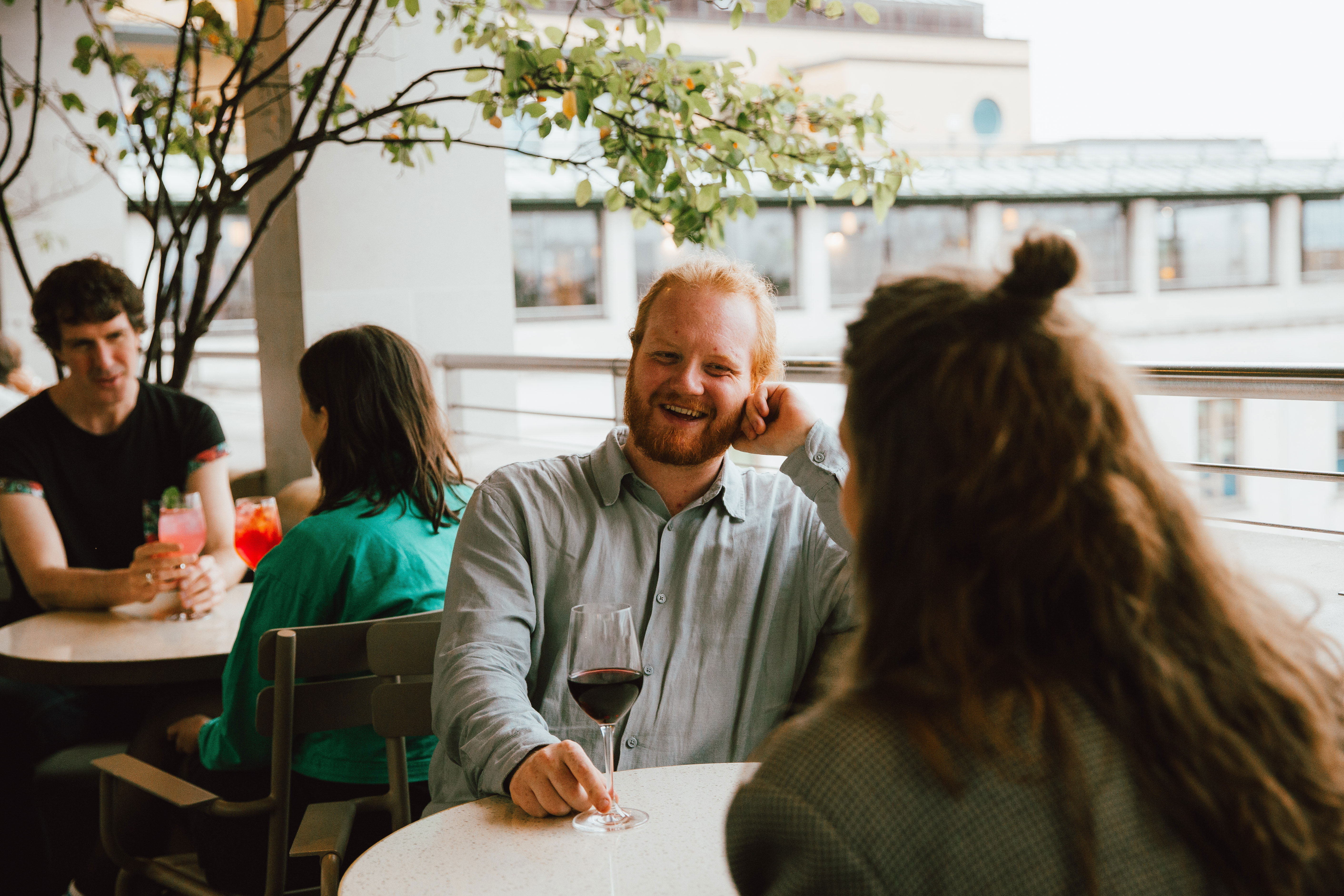 A group of people enjoys drinks on a terrace bar in Central London. A man with a beard and a wine glass smiles warmly, chatting with a woman who is facing away from the camera. 