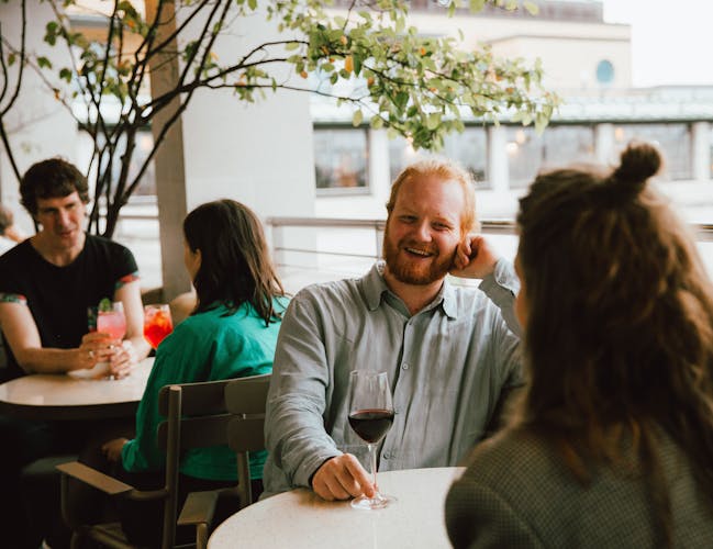 A group of people enjoys drinks on a terrace bar in Central London. A man with a beard and a wine glass smiles warmly, chatting with a woman who is facing away from the camera.