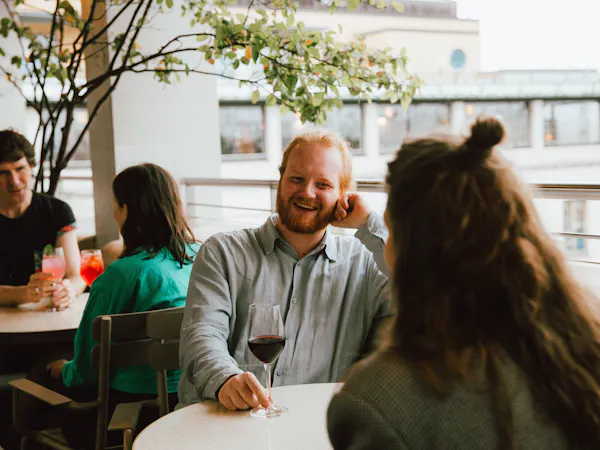 A group of people enjoys drinks on a terrace bar in Central London. A man with a beard and a wine glass smiles warmly, chatting with a woman who is facing away from the camera.