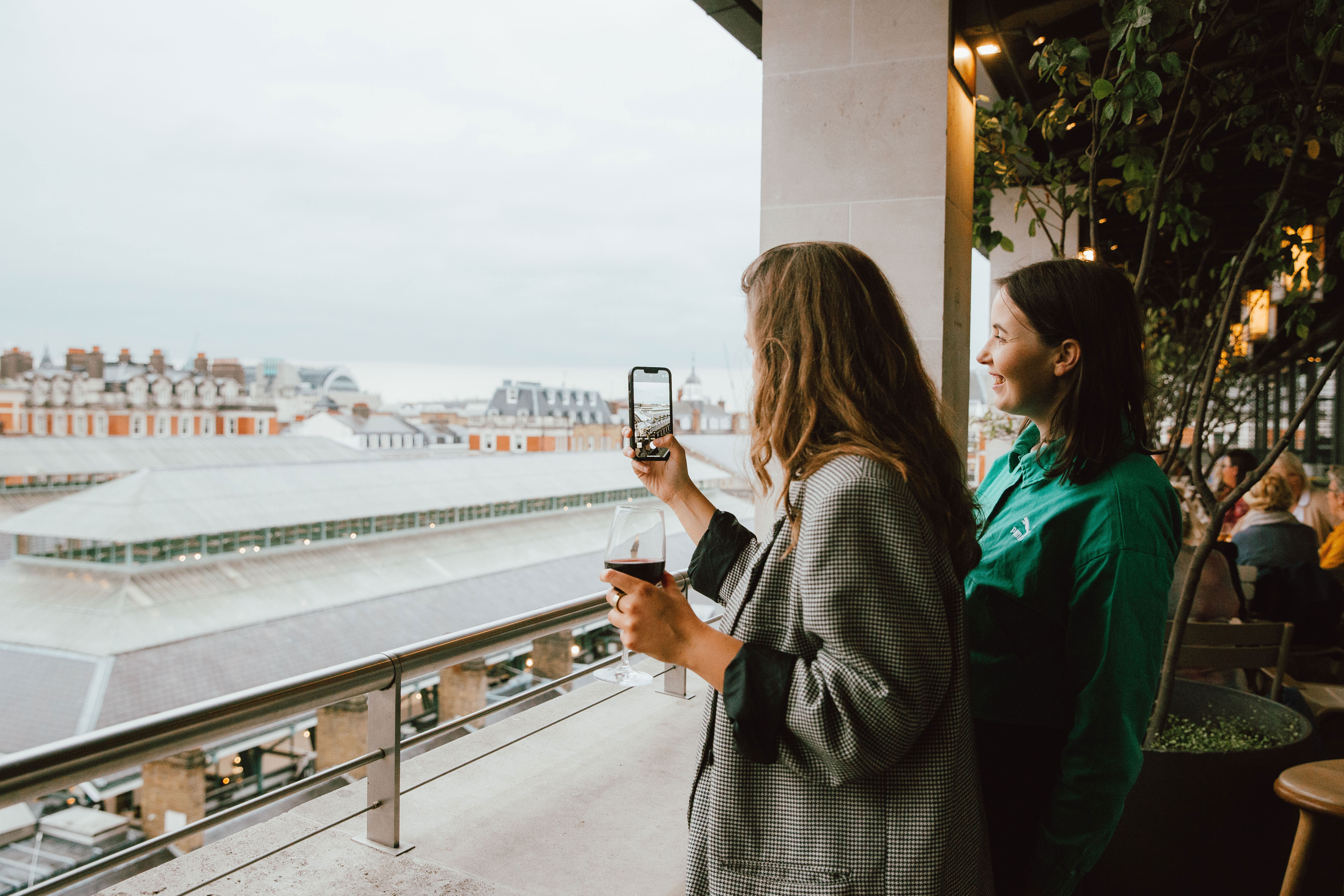 Two women on a rooftop terrace enjoy a cityscape view. One holds a phone, capturing the scenery, while the other smiles. 