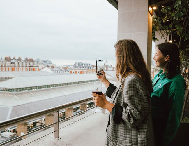 Two women on a rooftop terrace enjoy a cityscape view. One holds a phone, capturing the scenery, while the other smiles.