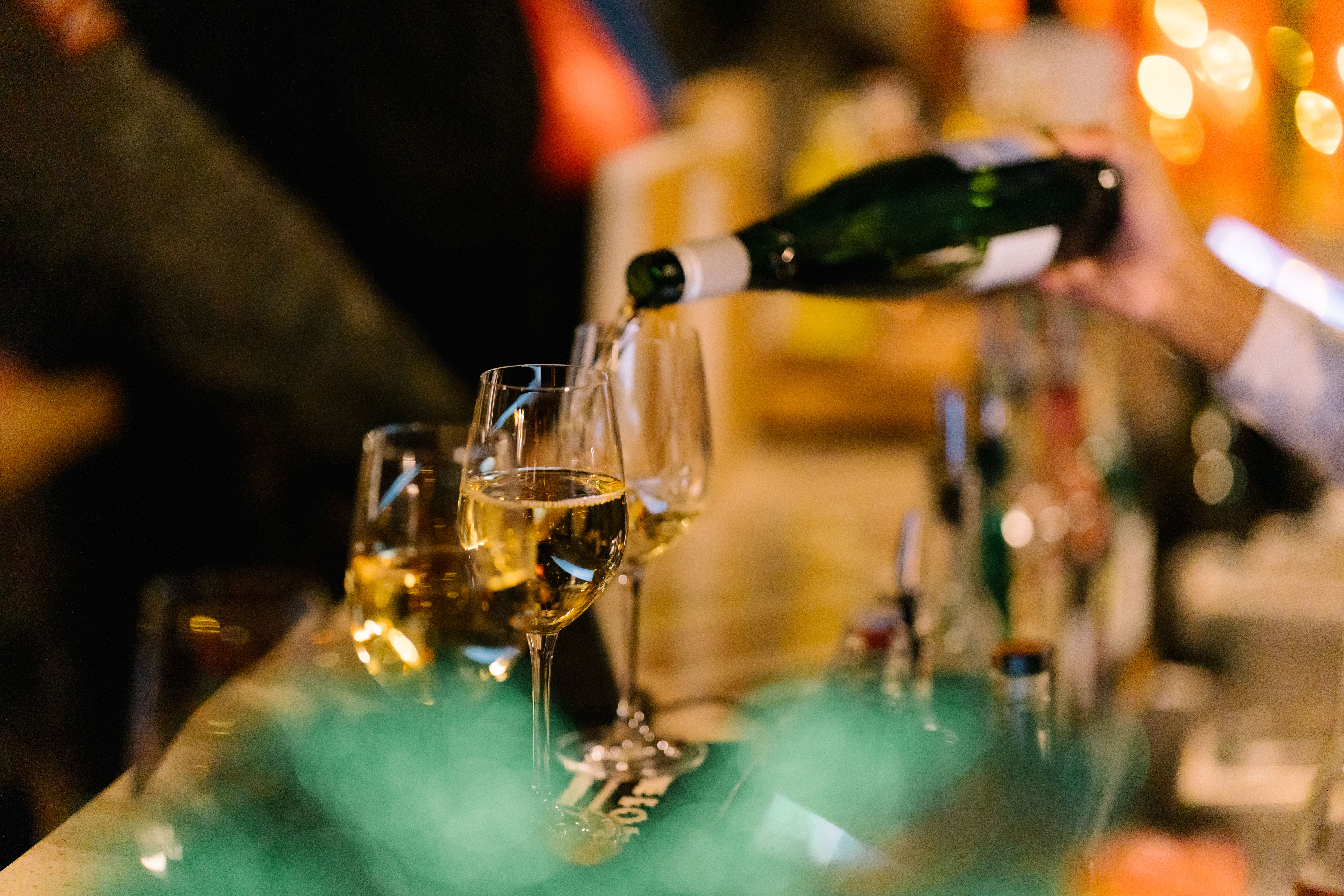 A hand pours white wine from a green bottle into a glass on a bar counter. The background features blurred warm lights. 