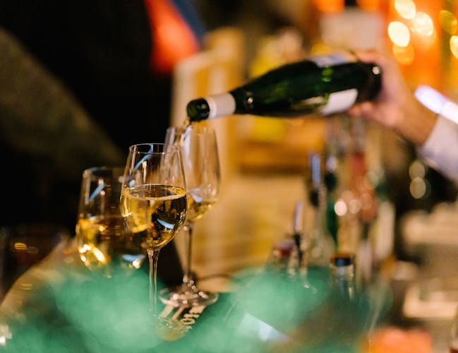 A hand pours white wine from a green bottle into a glass on a bar counter. The background features blurred warm lights.