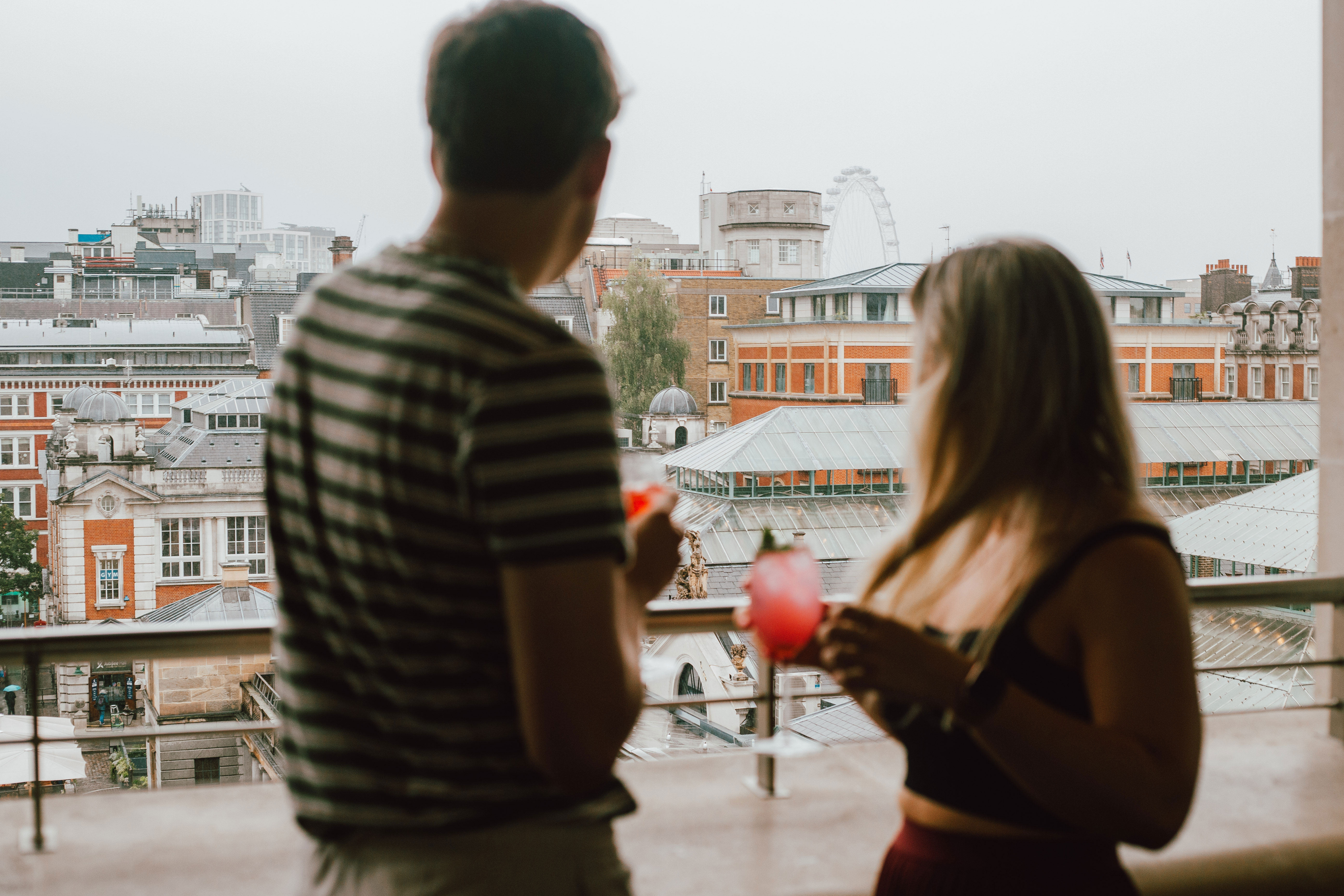 A man and woman holding drinks stand on a balcony overlooking an urban view with historic buildings and the London Eye ferris wheel on a foggy day.