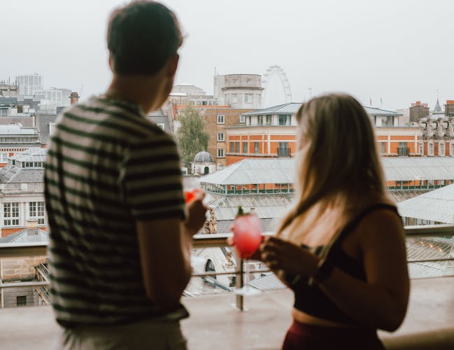 A man and woman holding drinks stand on a balcony overlooking an urban view with historic buildings and the London Eye ferris wheel on a foggy day.