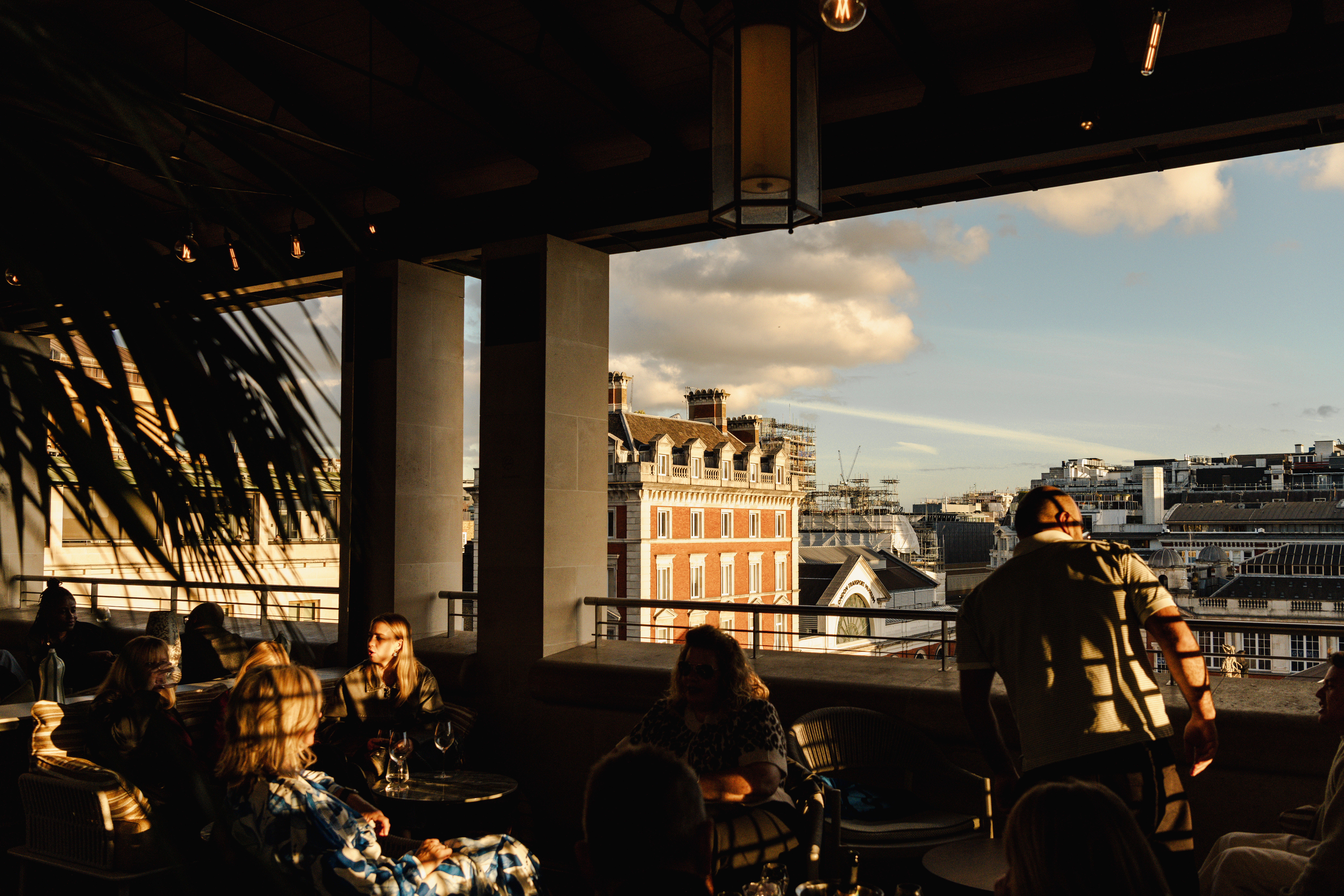 Sunlit rooftop terrace bar with people seated at tables, casting long shadows. A scenic city skyline is visible outside, under a partly cloudy sky.