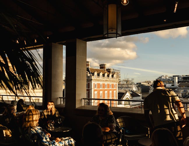 Sunlit rooftop terrace bar with people seated at tables, casting long shadows. A scenic city skyline is visible outside, under a partly cloudy sky.