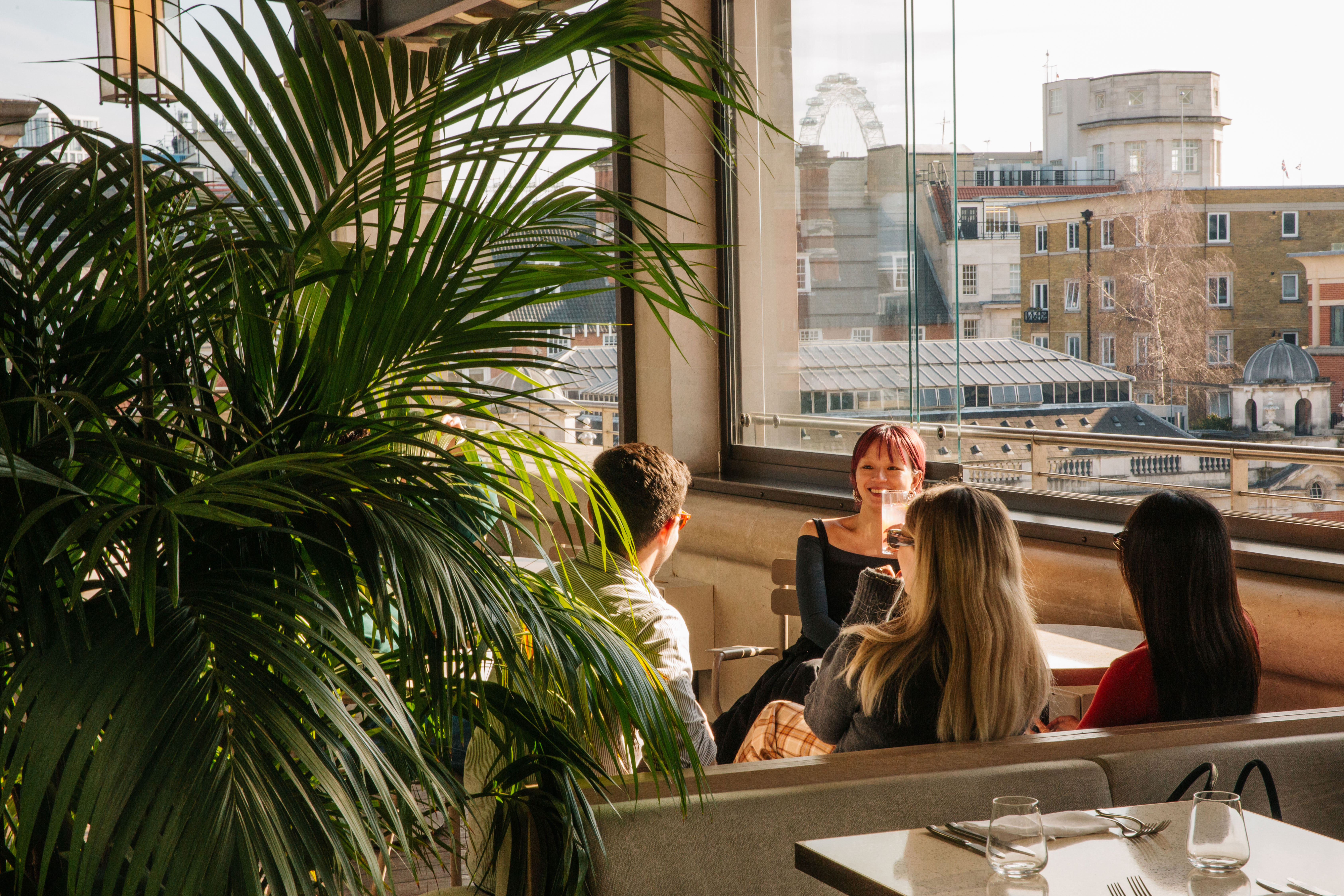 Four people sit around a table in a sunlit restaurant with large windows showcasing a central London city skyline. 