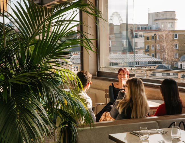 Four people sit around a table in a sunlit restaurant with large windows showcasing a central London city skyline.