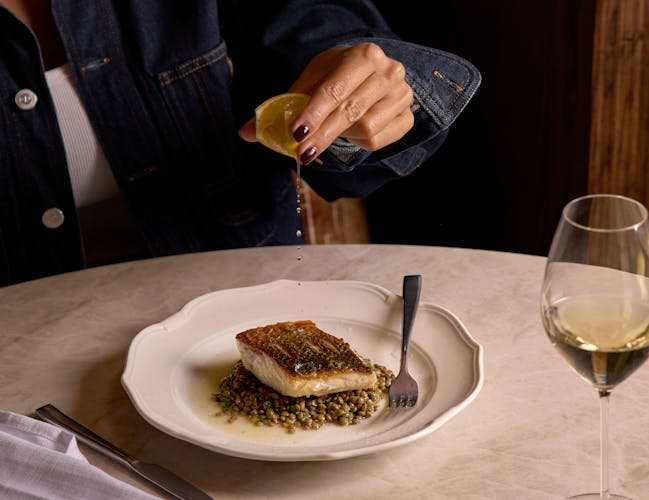 A person in a denim jacket squeezes a lemon over a plated fish dish with grains. A glass of white wine sits nearby on the white marble table.