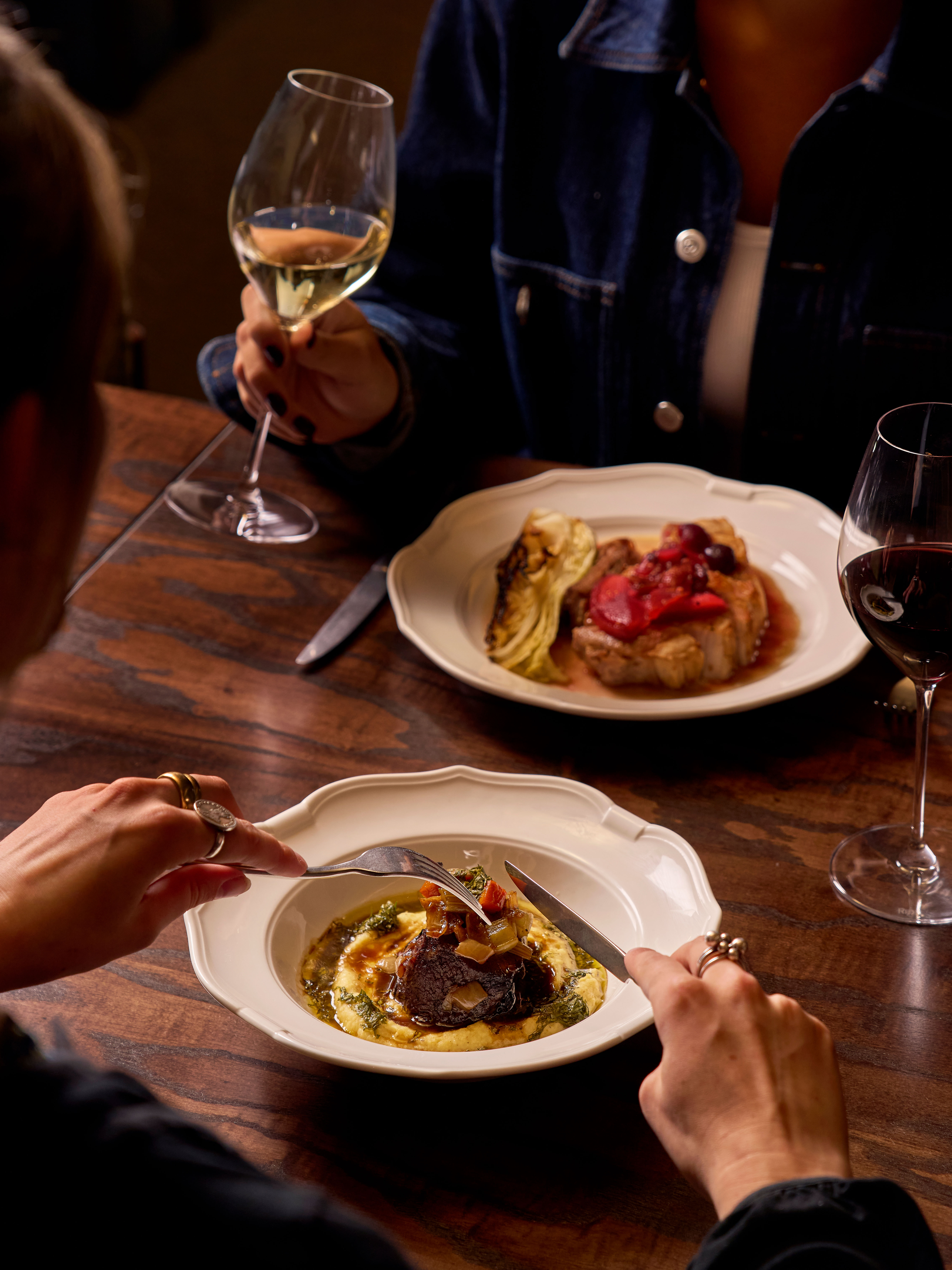 Two people dining at a wooden table, each with a glass of wine. One has a dish of meat and polenta, the other a plate with grilled meat and vegetables.
