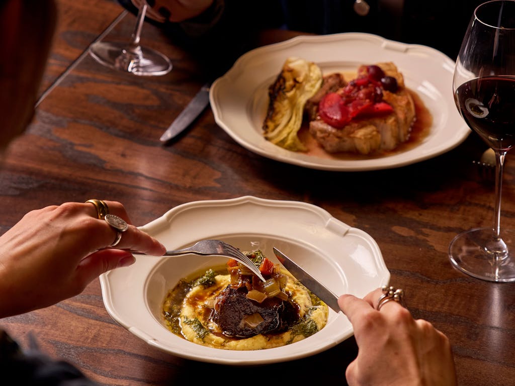 Two people dining at a wooden table, each with a glass of wine. One has a dish of meat and polenta, the other a plate with grilled meat and vegetables.