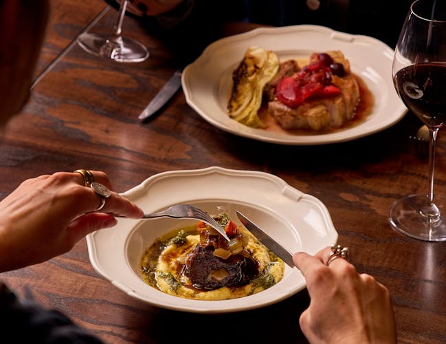 Two people dining at a wooden table, each with a glass of wine. One has a dish of meat and polenta, the other a plate with grilled meat and vegetables.