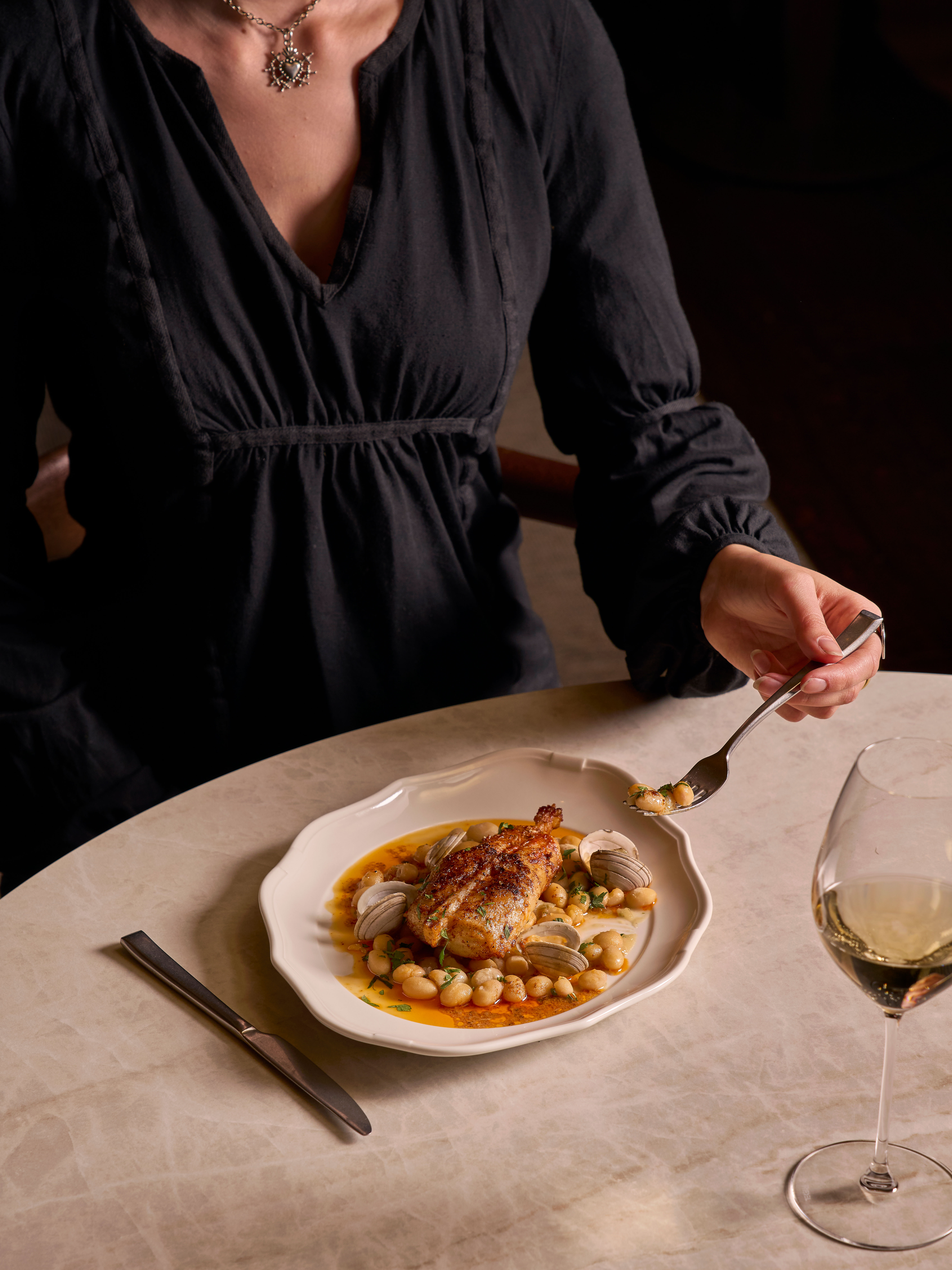 A person in a black shirt is seated at a marble table, holding a spoon over a dish of roasted chicken with clams and beans. A glass of white wine and cutlery are nearby. 