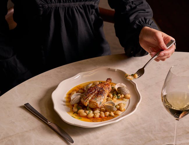 A person in a black shirt is seated at a marble table, holding a spoon over a dish of roasted chicken with clams and beans. A glass of white wine and cutlery are nearby.