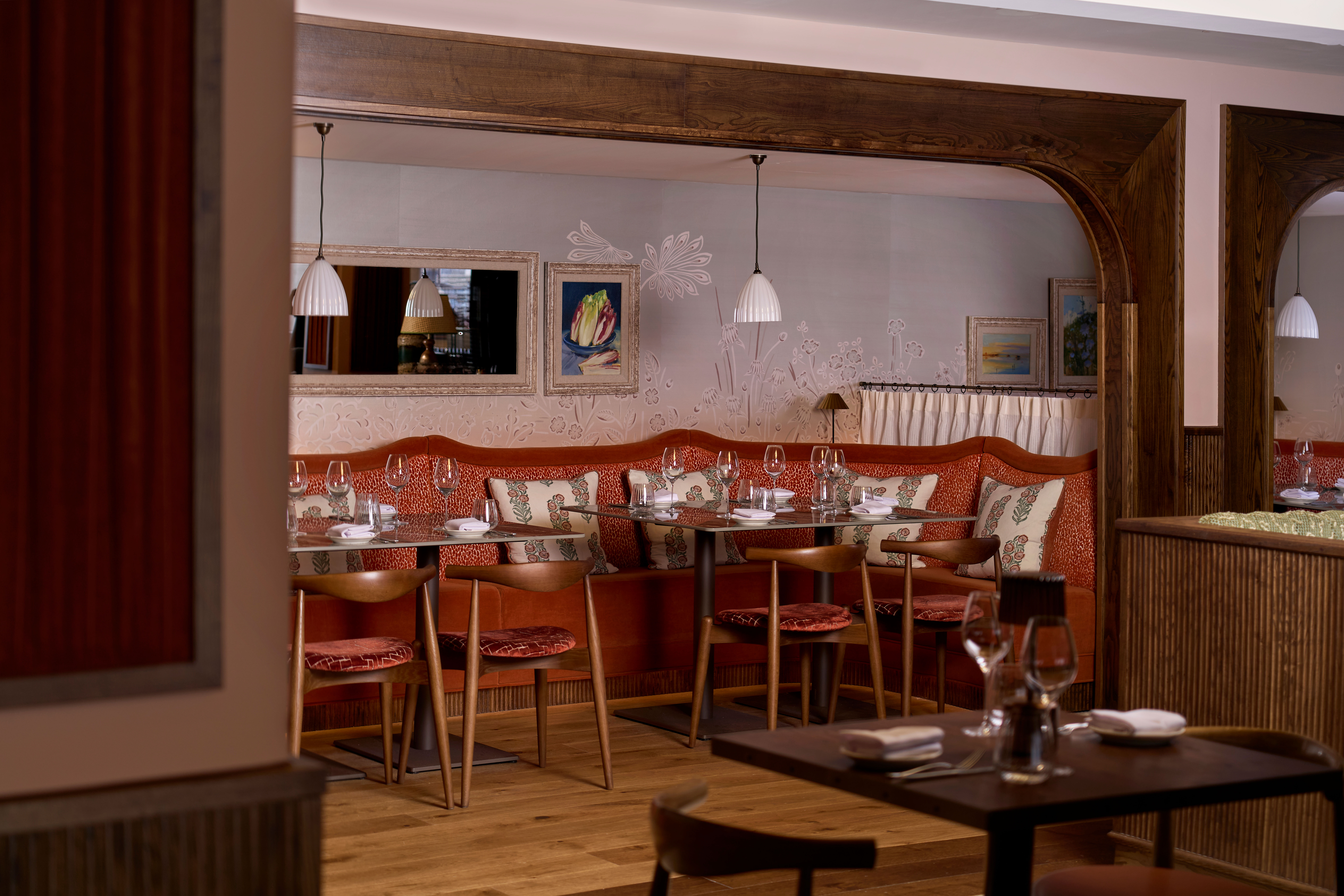 Cozy restaurant interior with red cushioned booths and wooden chairs. Elegant table settings with wine glasses. Warm lighting and art adorn the walls.