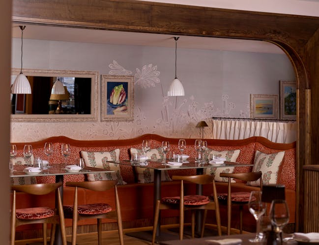 Cozy restaurant interior with red cushioned booths and wooden chairs. Elegant table settings with wine glasses. Warm lighting and art adorn the walls.