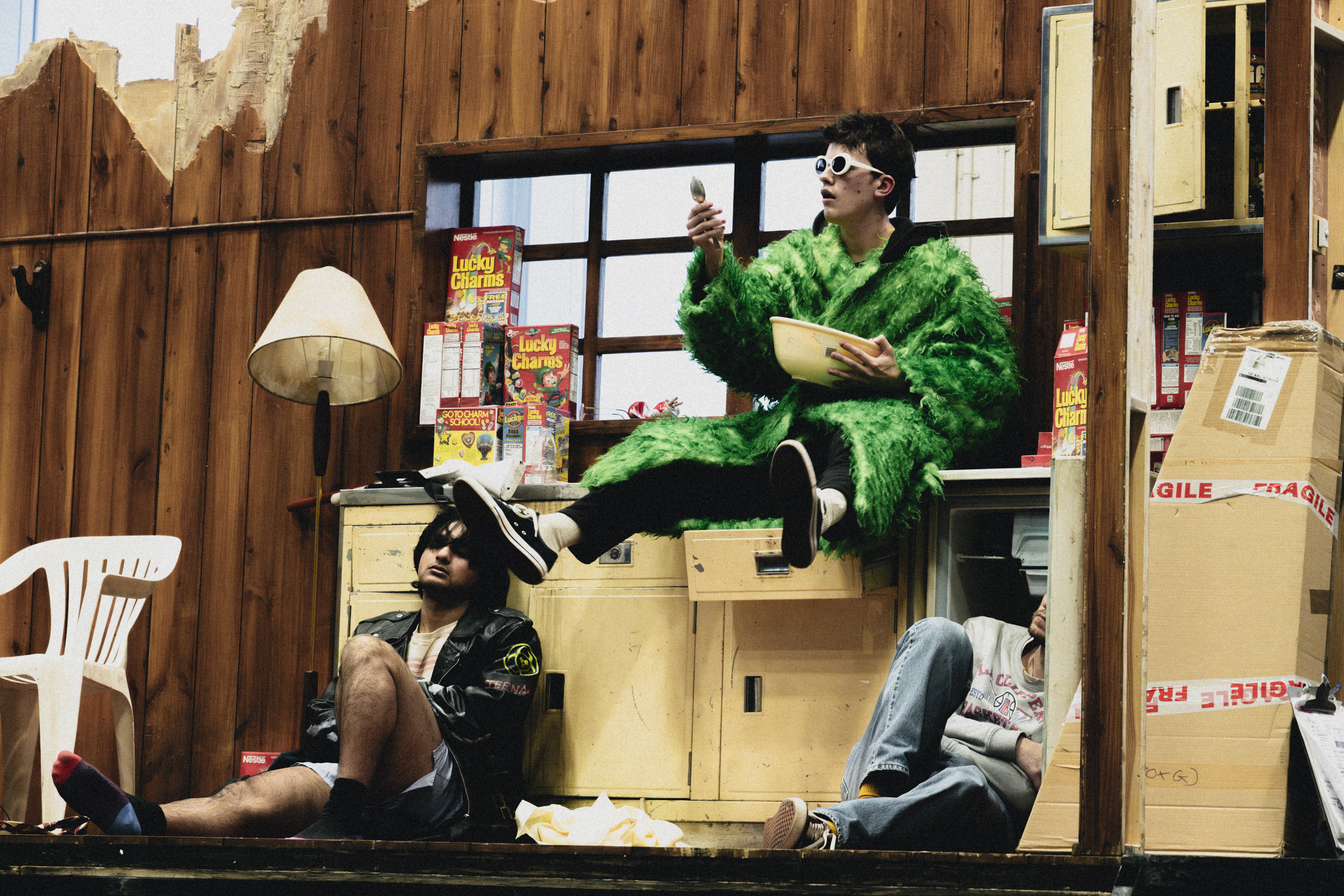 The stage is set to look like a neon-lit dirty kitchen filled with boxes of Lucky Charms cereal. A character wearing a green faux-fur coat, baggy taupe-coloured trousers, a pink baseball cap and white 60s style framed sunglasses sit on the counter pouring milk into a large bowl containing cereal. Below them sits another performer wearing a black puffer jacket, shorts, a black baseball cap and sunglasses. They are sitting against an open fridge. Another performer looks to be asleep on the floor with a blanket covering them over.