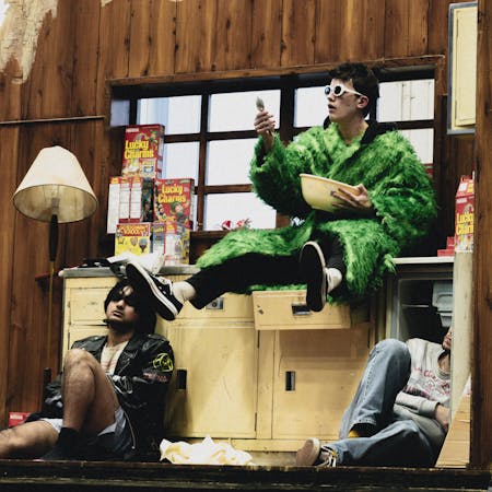 The stage is set to look like a neon-lit dirty kitchen filled with boxes of Lucky Charms cereal. A character wearing a green faux-fur coat, baggy taupe-coloured trousers, a pink baseball cap and white 60s style framed sunglasses sit on the counter pouring milk into a large bowl containing cereal. Below them sits another performer wearing a black puffer jacket, shorts, a black baseball cap and sunglasses. They are sitting against an open fridge. Another performer looks to be asleep on the floor with a blanket covering them over.