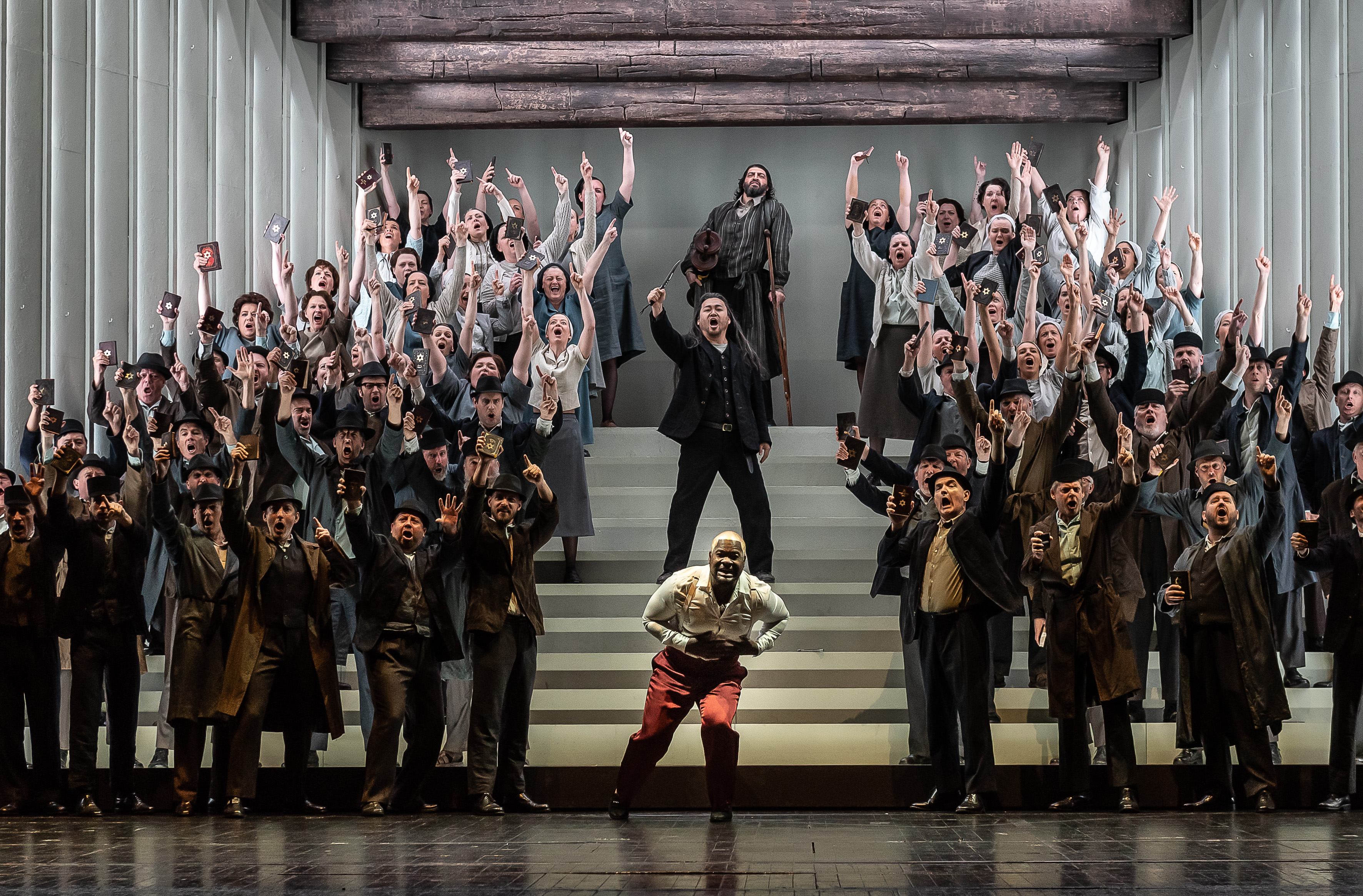 A chorus of singers stand on a stairs on a stage with their arms raised in the air, they are holding up a book with has a gold star embossed on the front. At the centre front, a character wearing red trousers lunges forward holding their chest. Behind them on the stairs leading up are two others, one has their right arm in the air holding up what looks like a knife. The character behind them leans on a crutch. 