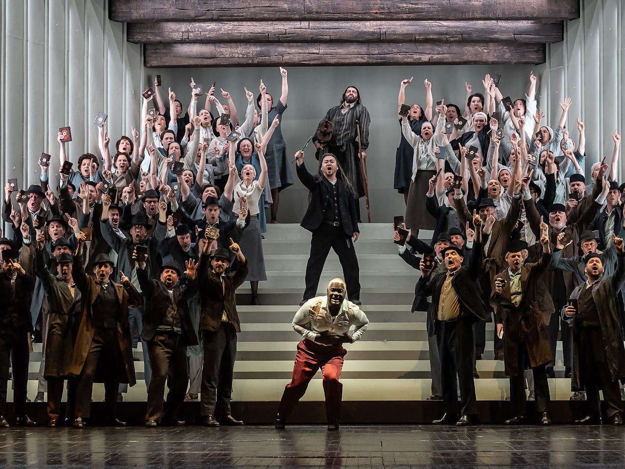 A chorus of singers stand on a stairs on a stage with their arms raised in the air, they are holding up a book with has a gold star embossed on the front. At the centre front, a character wearing red trousers lunges forward holding their chest. Behind them on the stairs leading up are two others, one has their right arm in the air holding up what looks like a knife. The character behind them leans on a crutch.