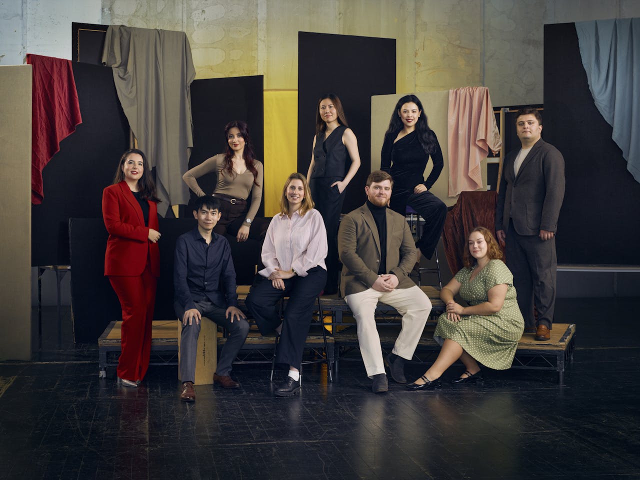 Nine people pose, either sitting down or standing, in a photography studio. Behind them different colour fabrics are draped over boards. They are all members of The Jette Parker Artists Programme.