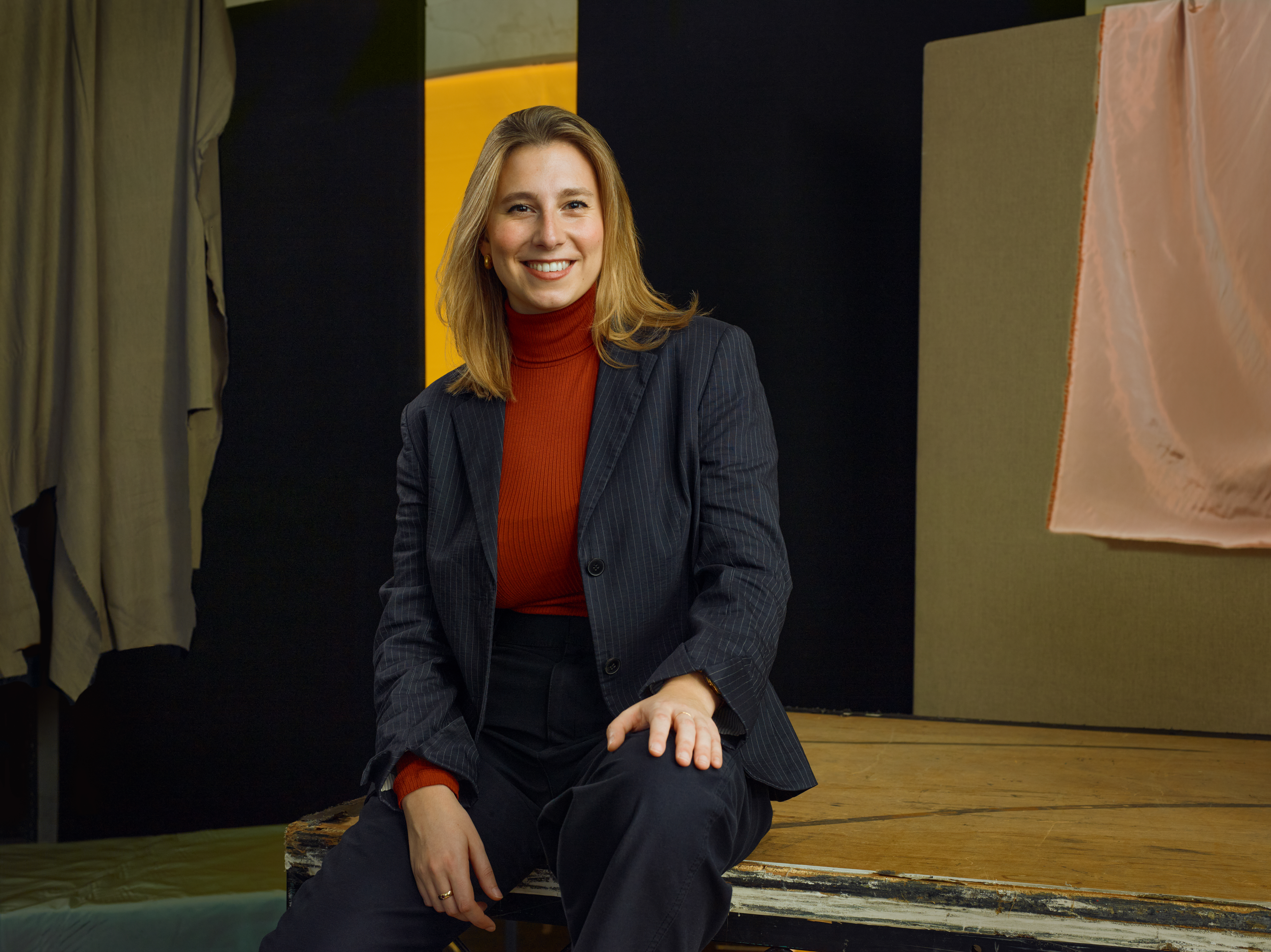 Jette Parker Artist Talia Stern sits in the centre of the photo, she is wearing a red turtle neck jumper and blue suit jacket and trousers.