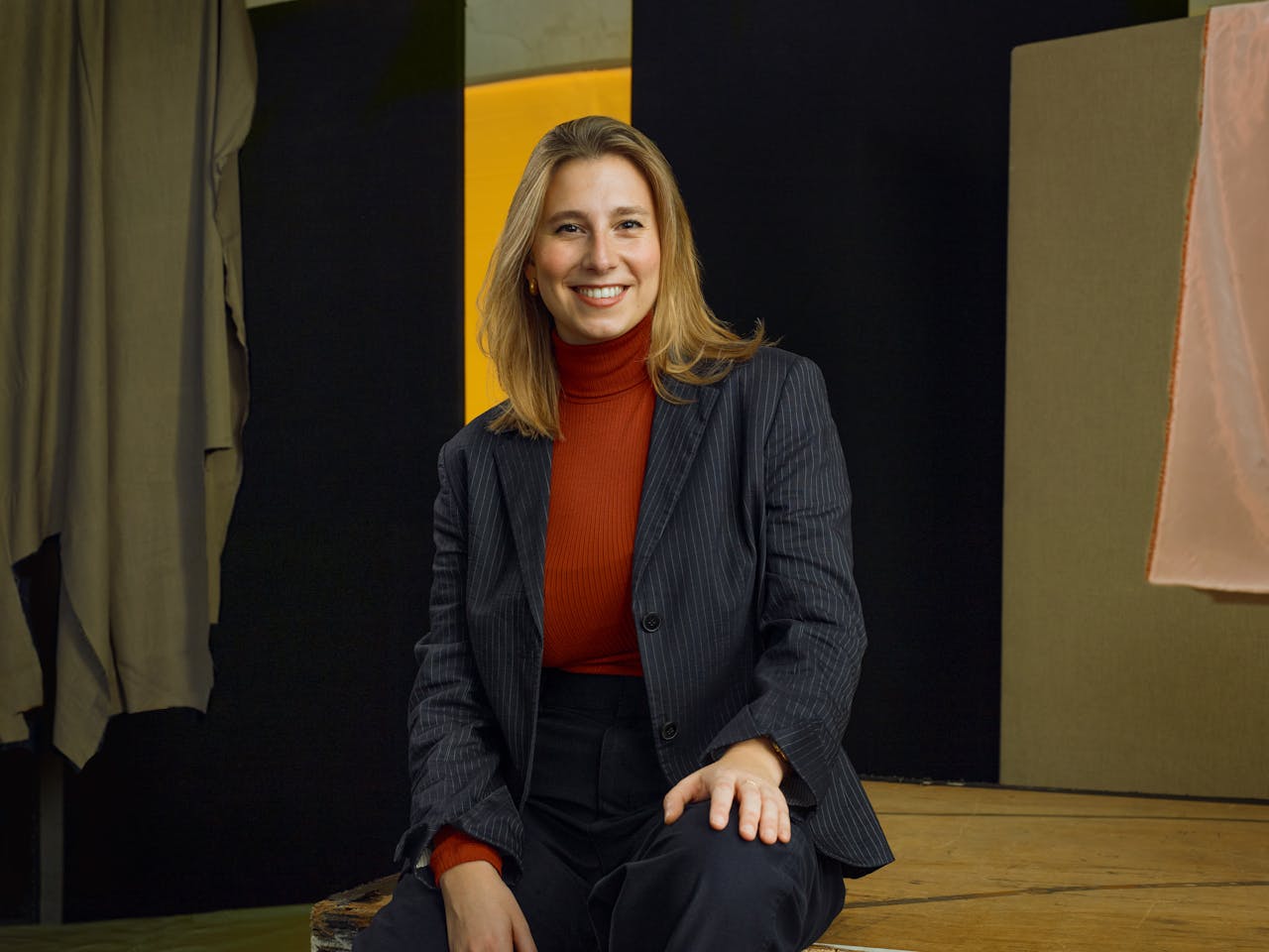 Jette Parker Artist Talia Stern sits in the centre of the photo, she is wearing a red turtle neck jumper and blue suit jacket and trousers.